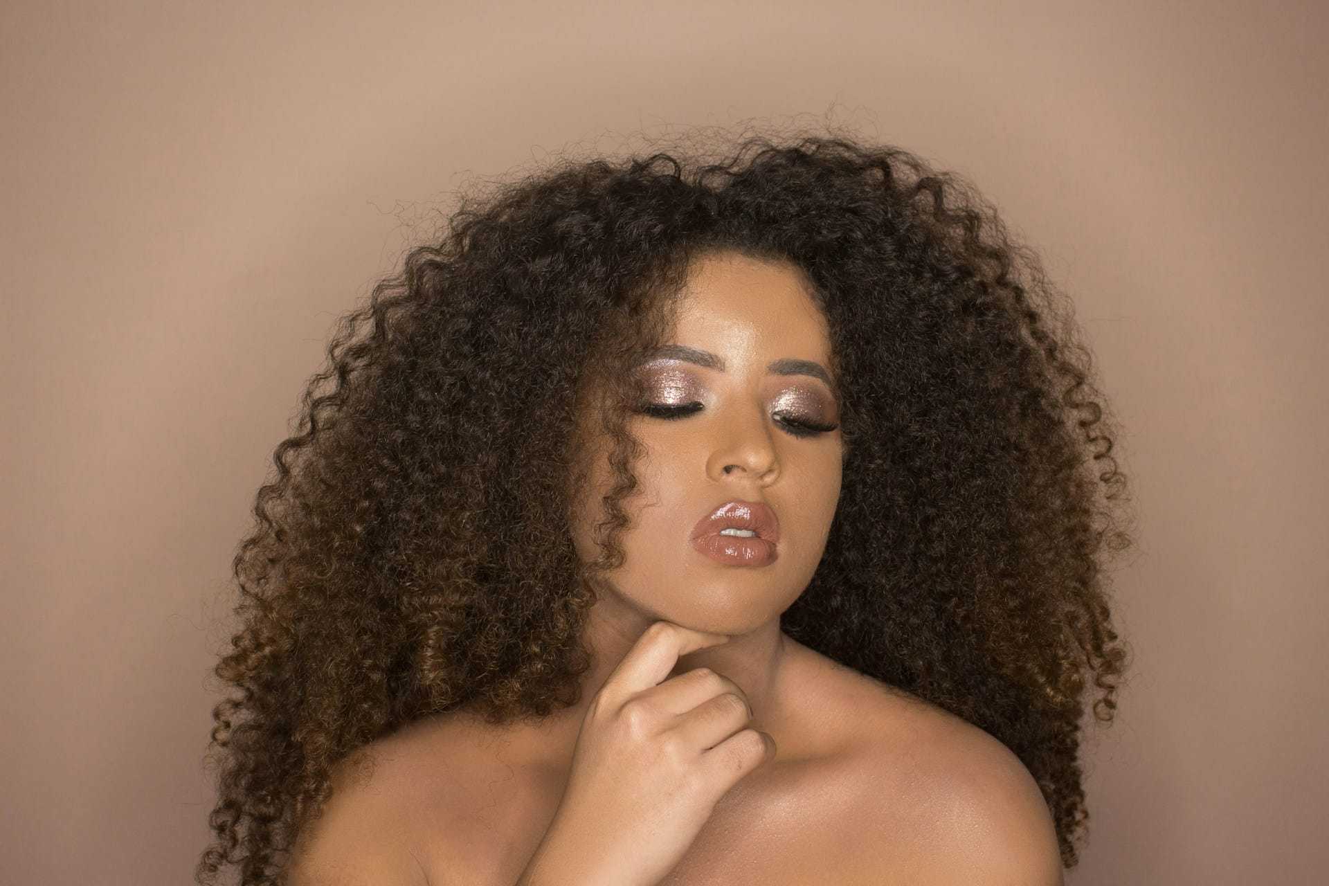 Woman with curly hair and shimmering makeup in thoughtful pose against a brown background.