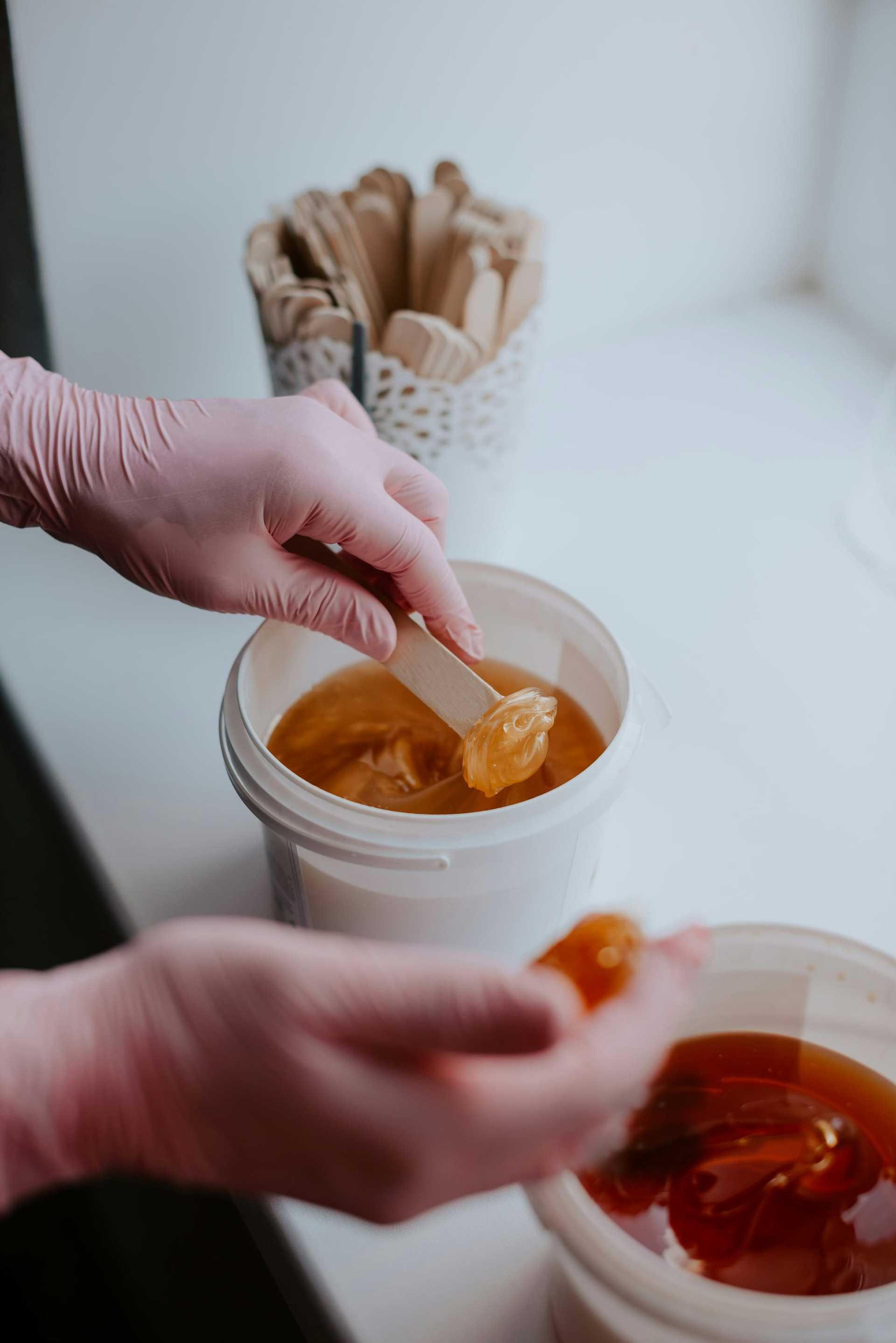 Hands in pink gloves applying wax from a container using wooden spatulas.