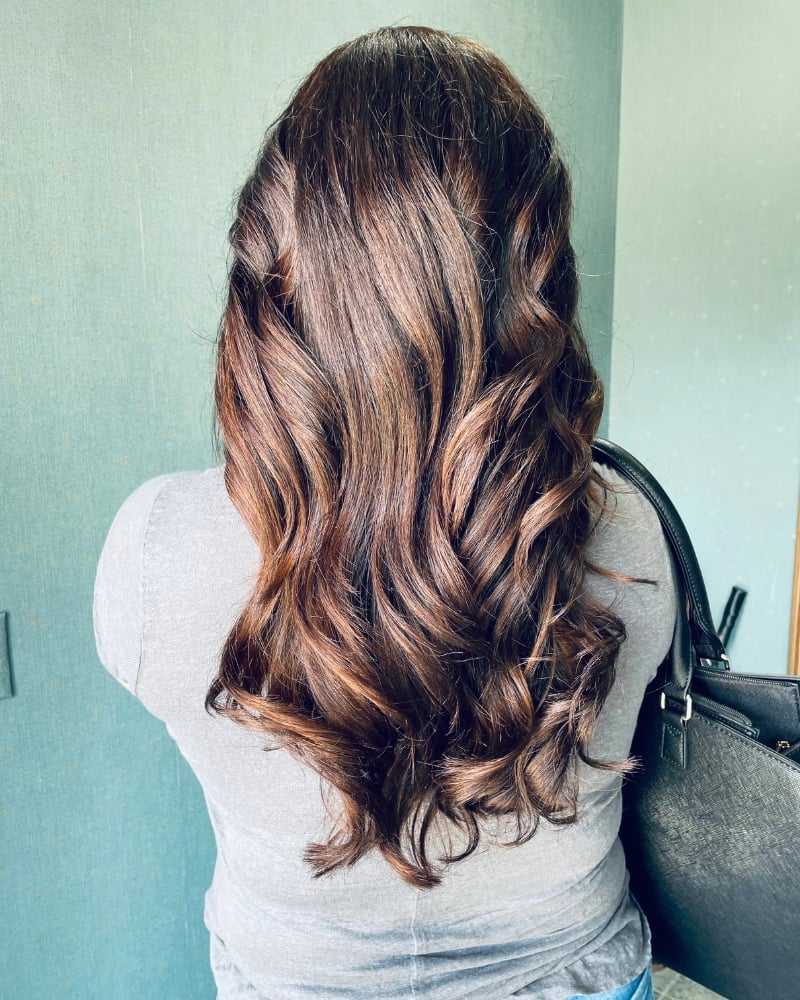 Long wavy brown hair styled on a woman's back, wearing a grey top and carrying a bag.